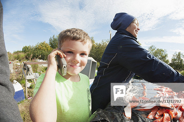 Ein Junge telefoniert mit einem Handy  während Erwachsene gefangenen Lachs filetieren und aufhängen  Bristol Bay  Alaska