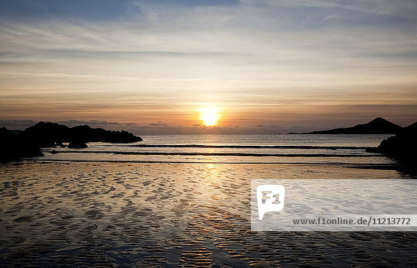 'Sun setting over the horizon with the water washing onto rippled sand on the shore and silhouetted rocks along the coast; Castlecove County Kerry Ireland'