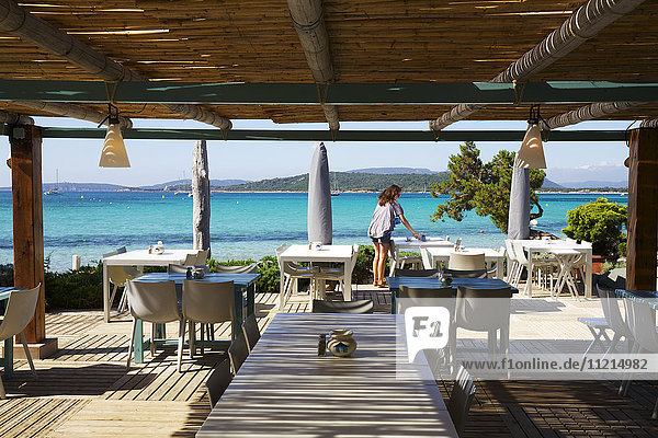Kellnerin beim Aufstellen der Tische in einem Strandcafé mit türkisblauem Meerblick