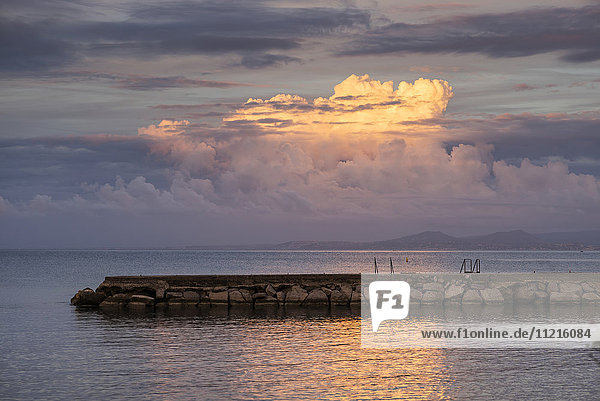 'A stone pier out in the tranquil water reflecting the glowing clouds at sunset over the mediterranean; Lacco Ameno  Ischia  Naples  Campania  Italy'