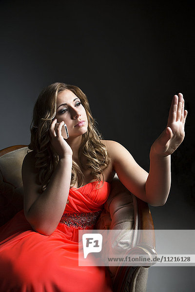 Eine schöne junge Frau sitzt in einem orangefarbenen Kleid auf einem Stuhl und betrachtet ihre Nägel  während sie mit ihrem Smartphone in einem dunkel beleuchteten Studio mit weißem Hintergrund telefoniert; Edmonton  Alberta  Kanada'.