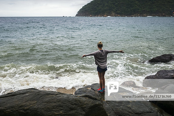 Eine junge Frau steht auf einem Felsen am Wasser und blickt mit ausgestreckten Armen auf den Ozean; Yalepa  Jalisco  Mexiko'.