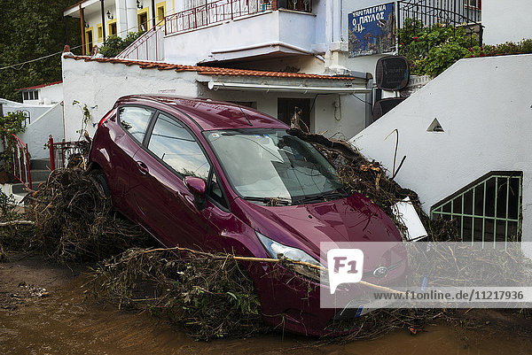 An abandoned car outside a house sloped onto dead trees laying in the street; Skopelos Greece