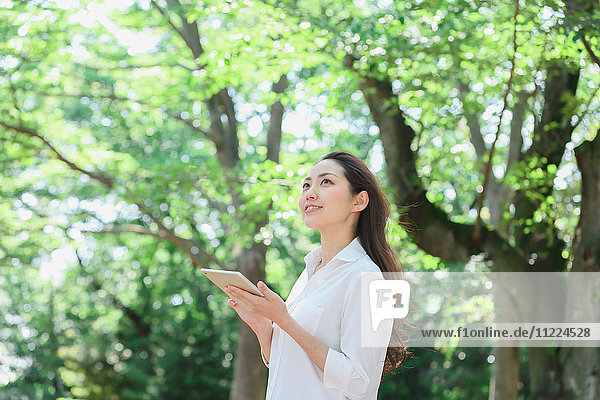 Junge Japanerin mit Tablet inmitten von Grün in einem Stadtpark
