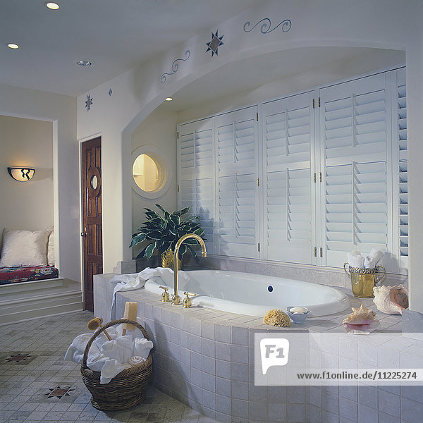 Badewanne mit Flieseneinfassung in gewölbter Nische mit Fensterläden  rundes Fenster. Korb mit Handtüchern auf gefliestem Boden  südwestliche Muster  rundes Fenster
