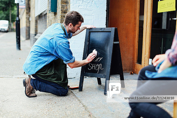 Junger Mann kniet auf dem Bürgersteig und wischt die Tafel vor der Werkstatt
