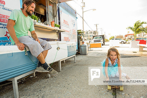 Vater sitzt neben Fastfood-Anhänger und beobachtet Tochter auf Skateboard