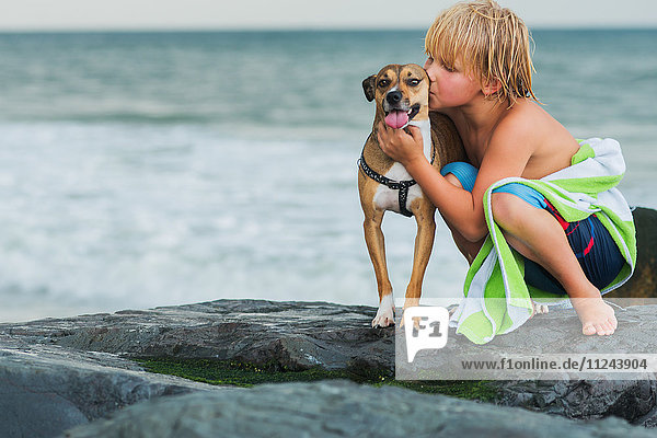 Young boy crouching on rocks at beach  hugging pet dog