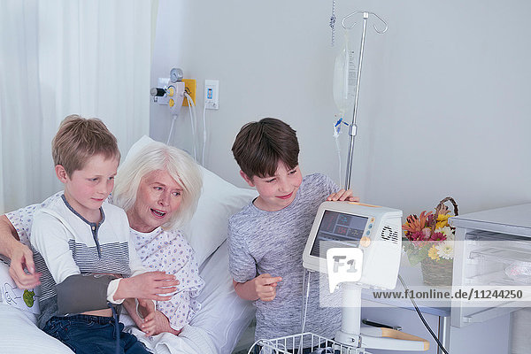 Ältere Patientin im Krankenhausbett mit Enkeln  die mit einem Blutdruckmessgerät spielen