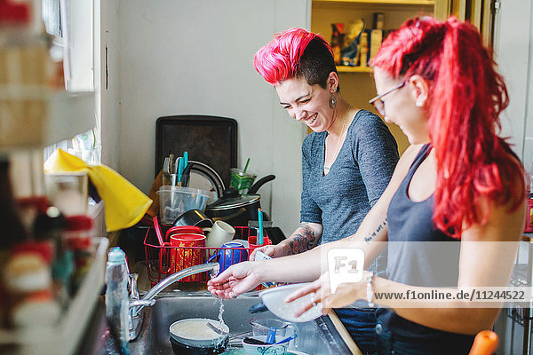 Zwei junge Frauen mit rosa Haaren lachen beim Geschirrspülen an der Spüle