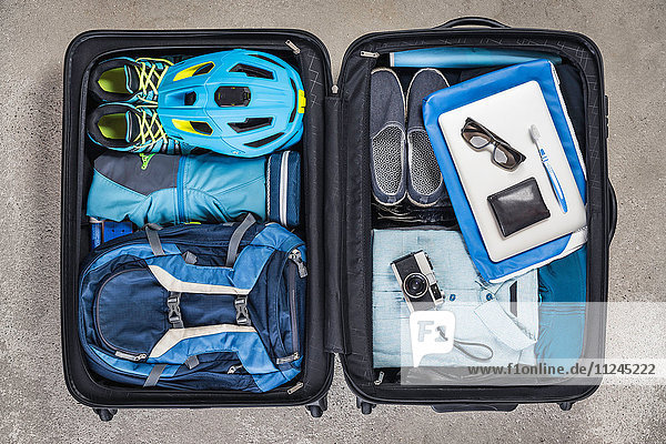 Draufsicht auf offenen gepackten Koffer mit blauem Hemd  Fahrradhelm  Rucksack  Retro-Kamera und Zahnbürste