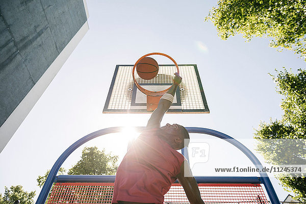 Junger männlicher Basketballspieler wirft Ball im sonnenbeschienenen Basketballkorb