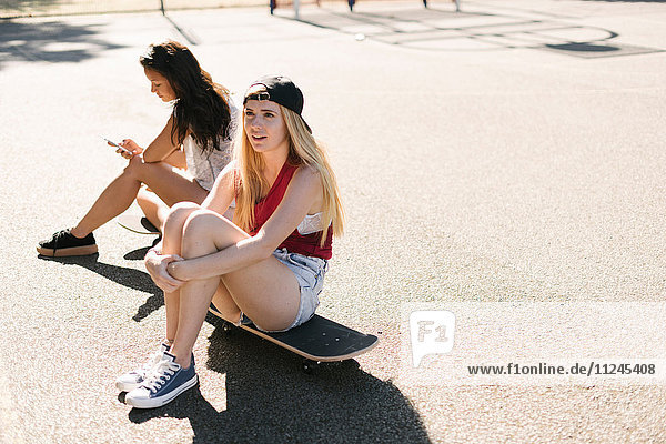 Two adult female friends sitting on skateboards on basketball court