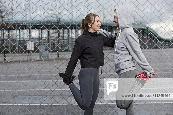 Two female runner friends stretching legs by wire fence