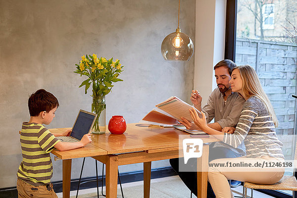 Mid adult couple looking at paperwork on dining table whilst son uses laptop