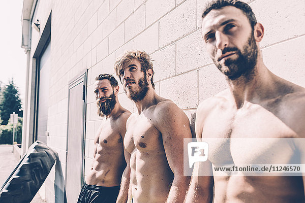 Drei männliche Kreuztrainer mit nacktem Oberkörper  die sich vor der Turnhalle an die Wand lehnen