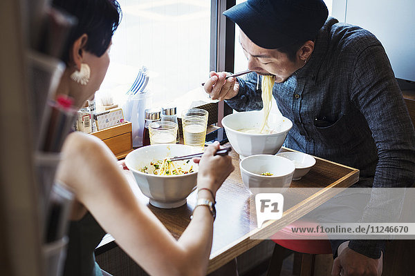 Ein Ramen-Nudel-Café in einer Stadt. Ein Mann und eine Frau sitzen und essen Nudeln aus großen weißen Schalen.