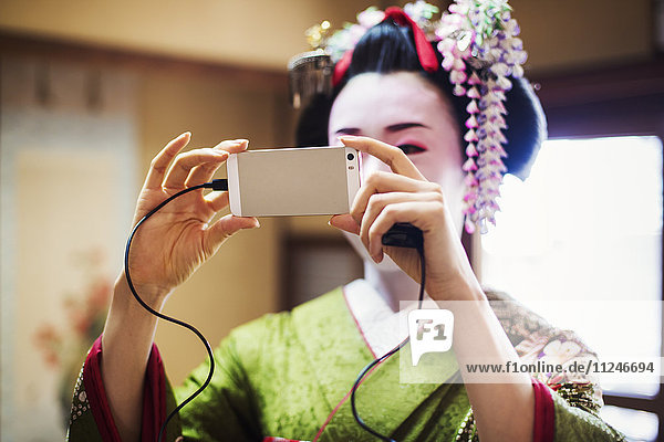 Eine im traditionellen Geisha-Stil gekleidete Frau in Kimono und Obi  mit aufwändiger Frisur und blumigen Haarspangen  mit weißer Gesichtsschminke mit leuchtend roten Lippen und dunklen Augen. Sie nimmt ein Selfie.