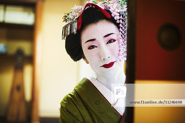 A woman dressed in the traditional geisha style  wearing a kimono and obi  with an elaborate hairstyle and floral hair clips  with white face makeup with bright red lips and dark eyes  looking in the mirror.