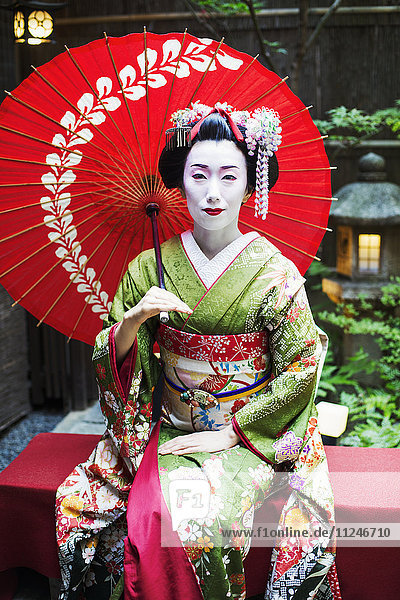 Eine im traditionellen Geisha-Stil gekleidete Frau in Kimono und Obi  mit aufwändiger Frisur und blumigen Haarspangen  mit weißer Gesichtsschminke mit leuchtend roten Lippen und dunklen Augen  die einen Regenschirm hält.