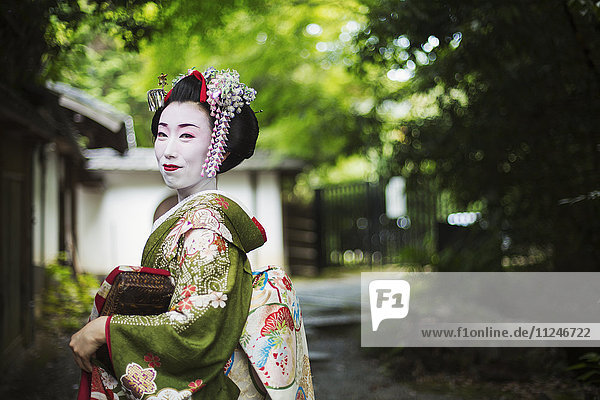 Eine im traditionellen Geisha-Stil gekleidete Frau in Kimono und Obi  mit aufwändiger Frisur und blumigen Haarspangen  mit weißer Gesichtsschminke mit leuchtend roten Lippen und dunklen Augen  Seitenansicht im Freien.