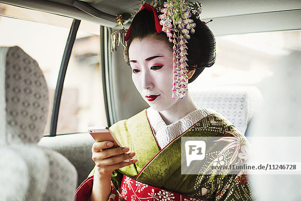 A woman dressed in the traditional geisha style  wearing a kimono and obi  with an elaborate hairstyle and floral hair clips  with white face makeup with bright red lips and dark eyes in a car using a smart phone.