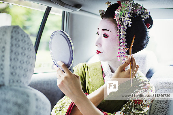 Eine im traditionellen Geisha-Stil gekleidete Frau in Kimono und Obi  mit aufwändiger Frisur und blumigen Haarspangen  mit weißer Gesichtsschminke mit leuchtend roten Lippen und dunklen Augen in einem Auto mit einem Handspiegel.