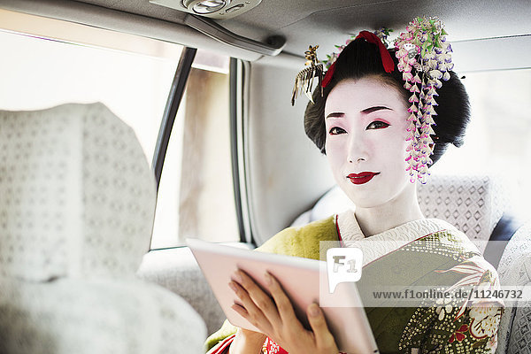 A woman dressed in the traditional geisha style  wearing a kimono with an elaborate hairstyle and floral hair clips  with white face makeup holding a digital tablet  travelling in a car.