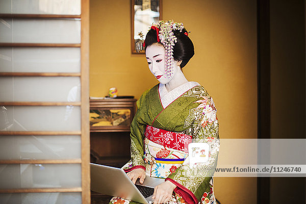 Eine im traditionellen Geisha-Stil gekleidete Frau in Kimono und Obi  mit aufwändiger Frisur und blumigen Haarspangen  mit weißem Gesichts-Make-up mit leuchtend roten Lippen und dunklen Augen  die einen Laptop-Computer benutzt.