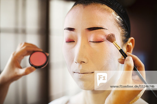 Eine Geisha oder Maiko mit einer Haar- und Make-up-Künstlerin  die die traditionelle Frisur und das Make-up mit einem Pinsel gestaltet  um Farbe auf ihre Augenlider aufzutragen.