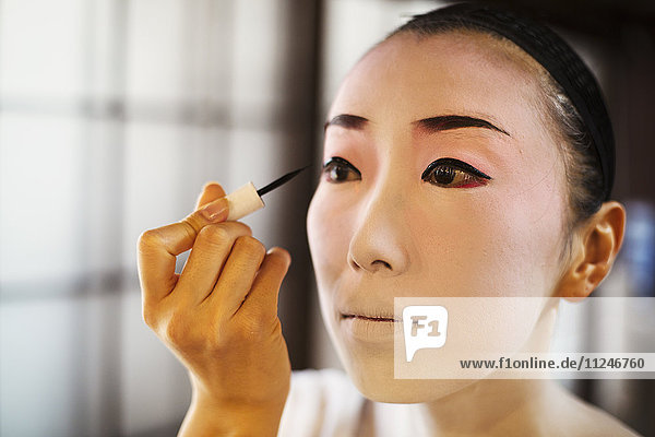 Geisha woman with traditional white face makeup painting on heavy eyeliner with a brush.