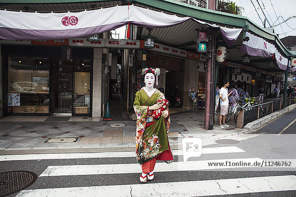 Eine im traditionellen Geisha-Stil gekleidete Frau  in Kimono und Obi  mit aufwändiger Frisur und blumigen Haarspangen  mit weißer Gesichtsschminke mit leuchtend roten Lippen und dunklen Augen  die eine Straße überquert.