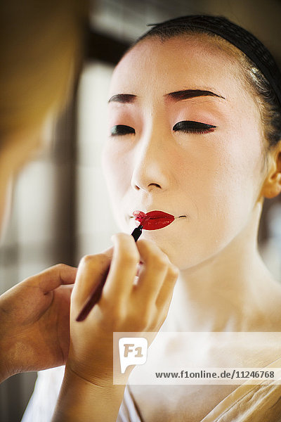 Eine moderne Frau  die mit einem feinen Pinsel auf Lippenstift die traditionellen lebendig roten Lippen der Geishas malt. Weißes Gesichts-Make-up.