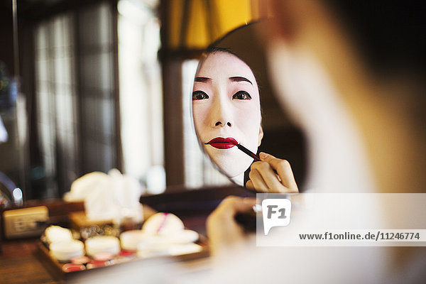 Geisha woman with traditional white face makeup applying bright red lipstick with a brush looking into a mirror.
