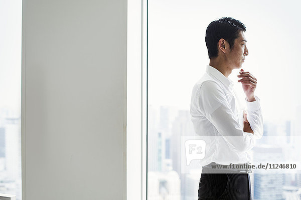 A businessman in the office standing by a large window with arms folded  looking over the city.
