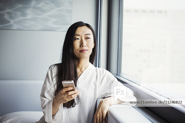 Eine Geschäftsfrau  die sich auf die Arbeit vorbereitet  in ihrer Nachtwäsche am Fenster sitzt und ein Smartphone in der Hand hält.