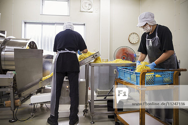 Arbeiterinnen und Arbeiter in Schürzen und Hüten sammeln frisch geschnittene Nudeln vom Förderband  um sie zu verpacken und zu verkaufen.