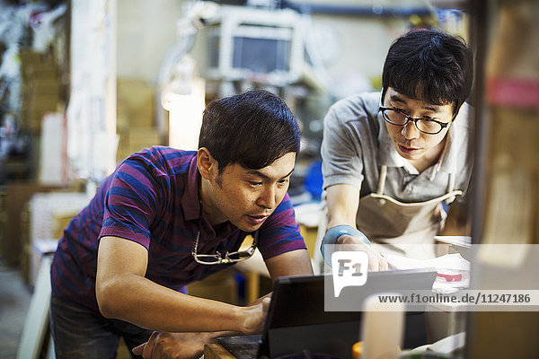 Two men using a laptop computer  working together in a glass maker's workshop.