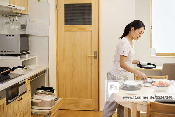 Familienhaus. Eine Frau  die in einer Küche eine Mahlzeit zubereitet und Geschirr auf den Tisch stellt.