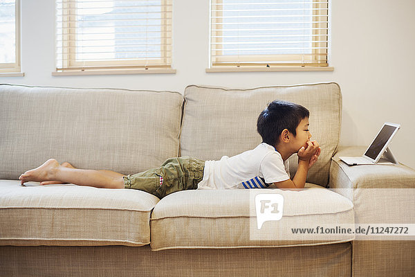 Familienhaus. Ein Junge liegt auf einem Sofa und betrachtet ein digitales Tablet.