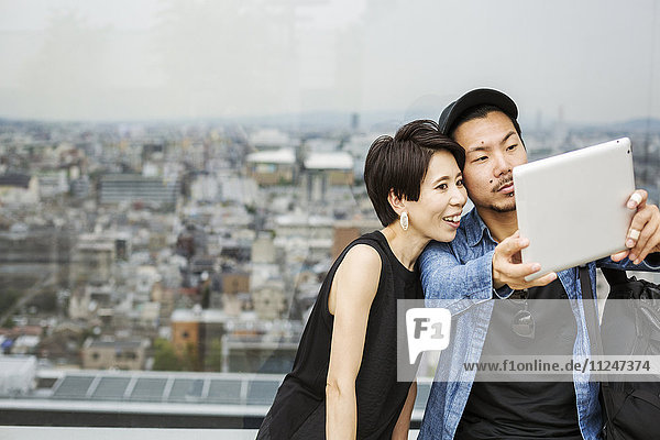 Zwei Menschen  ein Mann und eine Frau  die sich mit einem digitalen Tablett vor dem Blick über eine große Stadt ein Selfie machen.