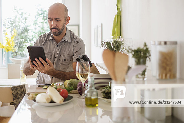 Mann mit digitalem Tablet an Küchentisch gelehnt
