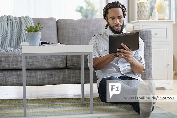 Young man with dreadlocks using digital table by sofa in living room