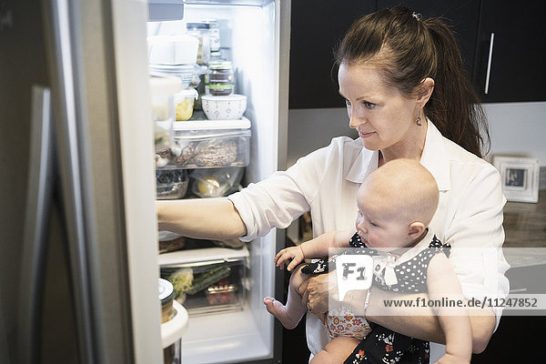 Mother looking for food in refrigerator while holding baby daughter (2-5 months)
