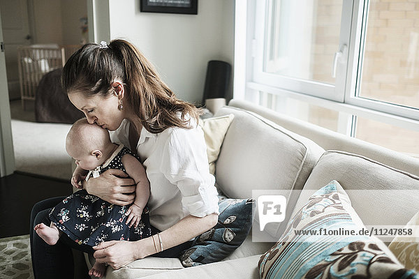Mutter küsst den Kopf ihrer kleinen Tochter (2-5 Monate) im Wohnzimmer