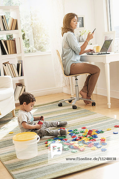 Boy (2-3) playing with toy blocks while mother working on laptop