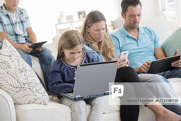 Family with two children (6-7  8-9) sitting on sofa  using laptop and digital tablets
