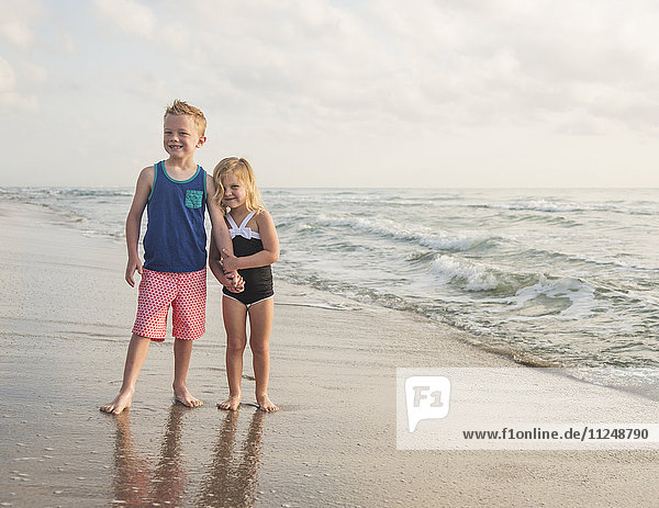 Junge (6-7) und Mädchen (4-5) stehen am Strand am Wasser und halten sich an den Händen
