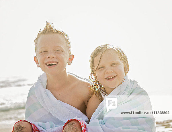 Junge (6-7) und Mädchen (4-5) sitzen in Handtücher eingewickelt am Strand