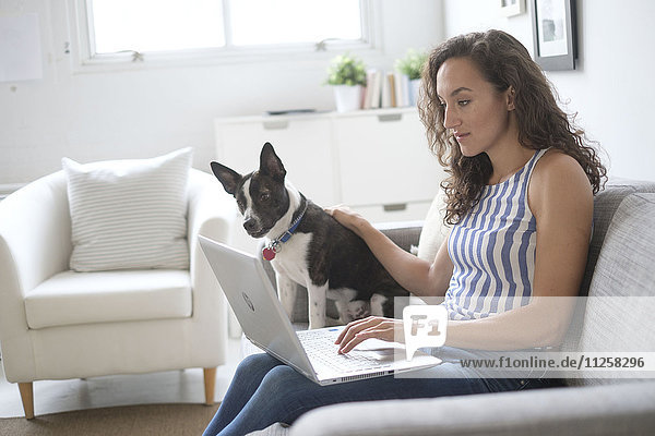 Junge Frau sitzt mit Laptop und Hund auf dem Sofa